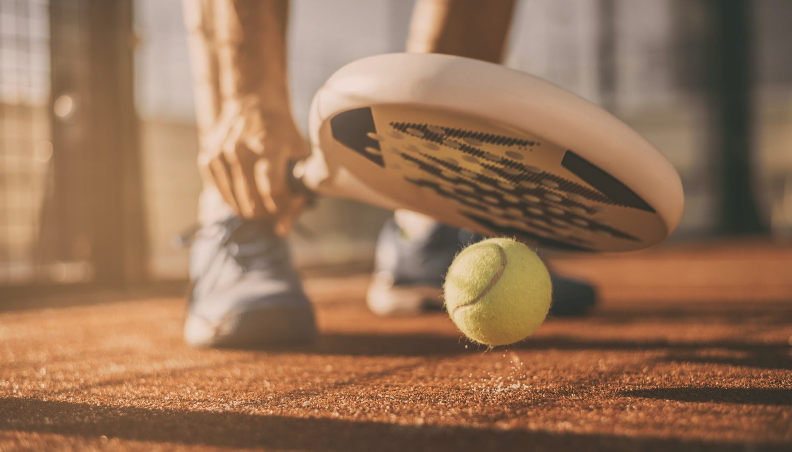 A padel racket lifting a ball off sunlit terracotta clay, court fencing behind